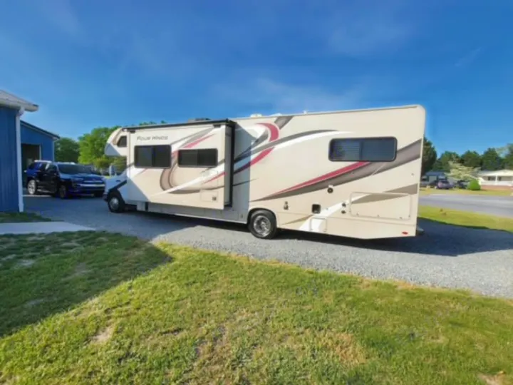 A light-colored RV rental, a Class C motorhome, parked on a gravel drive. Ready for your travel adventure under blue skies.. Thor Four Winds 2020