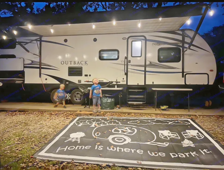 RV rental adventure: Two boys explore by a lit camper with a 'Home is where we park it' mat at night.. Keystone RV Outback Ultra-Lite 2018