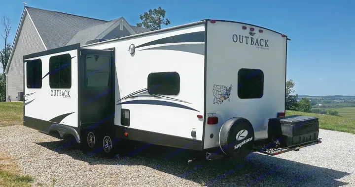 White Outback Ultra-Lite travel trailer RV rental with slide-out extended, parked on gravel with a scenic rural backdrop.. Keystone RV Outback Ultra-Lite 2018