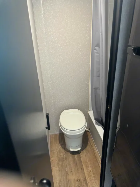 Compact RV rental bathroom features a white toilet, grey shower curtain, and wood-look floor.. Forest River Cherokee Wolf Pup Black Label 2022