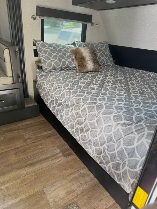 This RV rental interior features a cozy bed with stylish gray bedding and a window for scenic views, ideal for travel comfort.. Forest River Cherokee Wolf Pup Black Label 2022