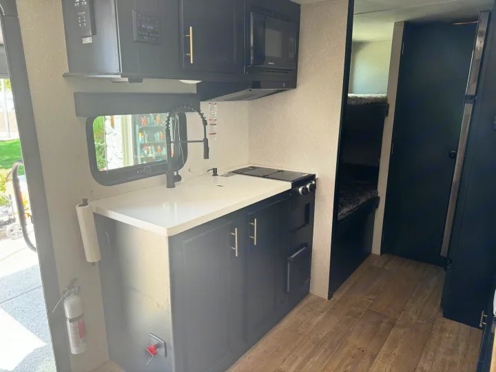 RV rental interior features a compact kitchen with a microwave, stove, and sink, plus a bunk bed sleeping area.. Forest River Cherokee Wolf Pup Black Label 2022