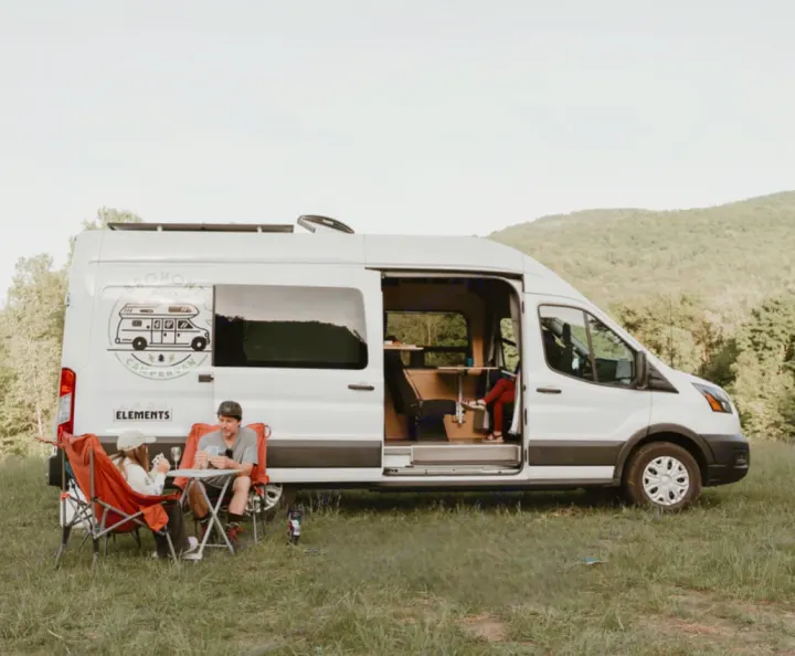 A couple enjoys cards by their **RV rental** camper van in a field. Perfect for a scenic outdoor escape! #rvrental #vanlife #camping. Ford Van 2023