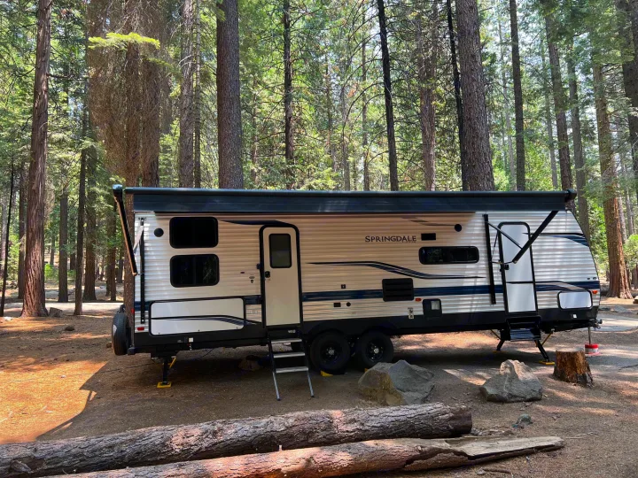 An RV rental parked among towering pine trees in a serene campground, ready for a forest retreat. Steps are down, awning out.. Keystone RV Springdale 2022