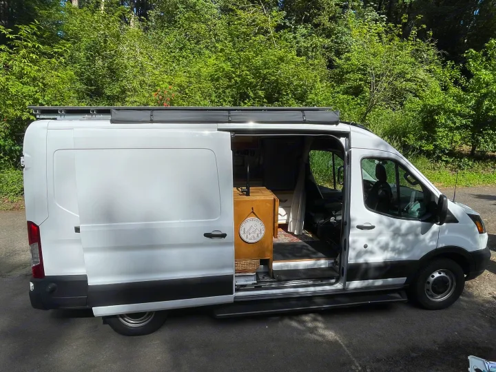 White high-top camper van with open door, showcasing custom wood interior for RV rental adventures. Includes sink, fridge, and 'Into the Woods' decor.. Ford Transit 2019