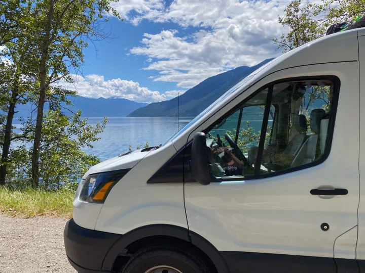 White camper van, an RV rental, parked on a dirt road by a serene lake with mountains and trees.. Ford Transit 2019