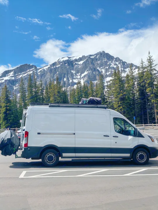 White RV rental van, equipped for adventure, parked before a stunning backdrop of snow-capped mountains and pine forests.. Ford Transit 2019