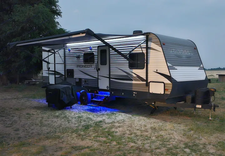 This illuminated RV rental, a Pioneer travel trailer, glows with blue underglow and awning lights at dusk, ideal for camping.. Heartland RVs Pioneer 2019