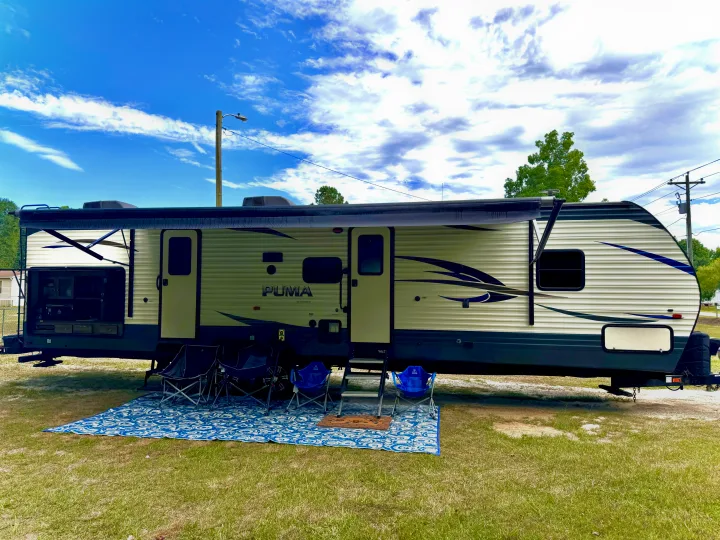 This Puma travel trailer RV rental, with awning, outdoor kitchen, mat, and chairs, is set up for camping on grass.. Forest River Palomino Puma 2018