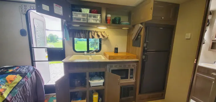 RV rental interior: A cozy, functional camper kitchen with sink, microwave, fridge, and storage. Open door view of nature.. Coachmen Apex Nano 2018