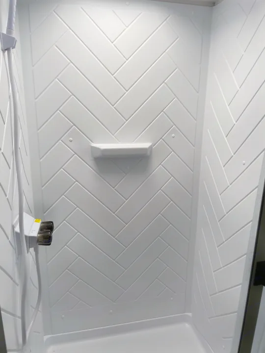 Clean RV rental shower featuring white herringbone pattern walls, shower controls, and an integrated shelf.. Forest River Wildwood X-Lite 2023