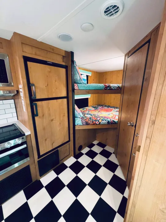 Inside an RV rental: Compact kitchen with Dometic fridge, Furrion stove, plus bunk beds and a bold checkered floor.. Riverside RV Retro 2023