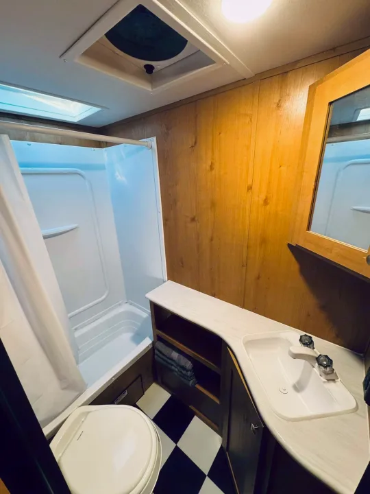 RV rental bathroom features: compact shower/tub, toilet, vanity with sink, mirror, wood walls, and checkerboard floor.. Riverside RV Retro 2023
