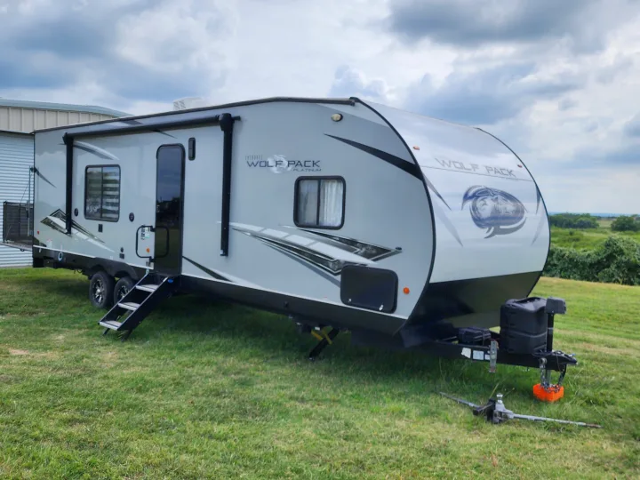 A grey Cherokee Wolf Pack Platinum RV rental, a travel trailer with steps down, parked on green grass under a cloudy sky.. Forest River Wolf Pack 2023