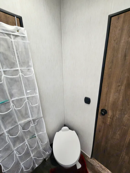 An RV rental bathroom with a white toilet, dark wood door, and a clear, over-the-door hanging organizer providing extra storage.. Forest River Wolf Pack 2023