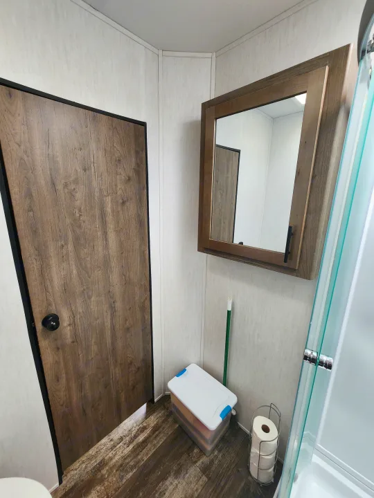 Inside an RV rental, showing a compact bathroom with a wood door, medicine cabinet, shower, and storage solutions.. Forest River Wolf Pack 2023
