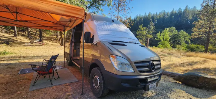 Golden RV rental Sprinter van with sunshade under an orange canopy at a serene, sunny forest campsite.. Mercedes-Benz Sprinter 2011