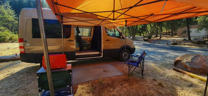 Tan Sprinter rv rental with an orange awning & open side for camp. Shows a blue chair, red cooler, and mat for outdoor living.. Mercedes-Benz Sprinter 2011