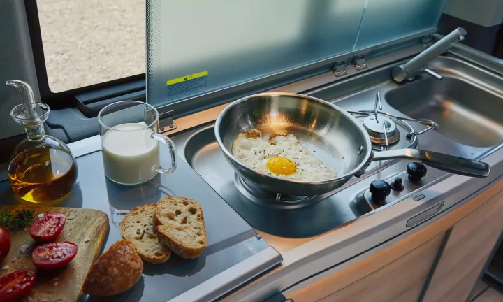 Compact rv rental kitchen prep: a fried egg cooks on the stove beside a sink. Fresh tomatoes, bread, milk, and oil are on the counter.. Volkswagen California T6.1 4MO 2024
