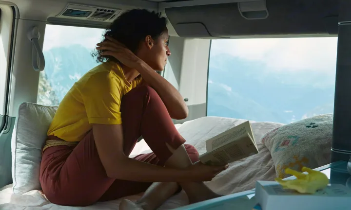 A woman enjoys her RV rental, reading a book in a cozy interior with stunning mountain views.. Volkswagen California T6.1 4MO 2024