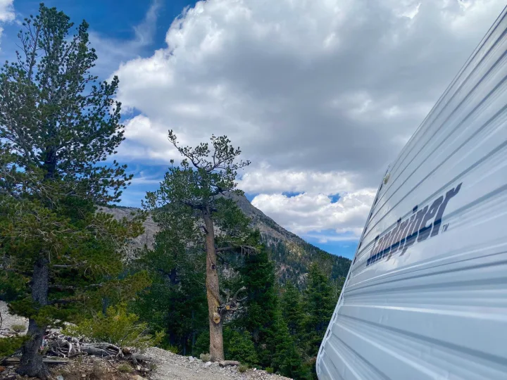 An RV rental is seen from a low angle, parked on a dirt path with pine trees and mountains under a cloudy sky.. Forest River Cherokee Lite 2000