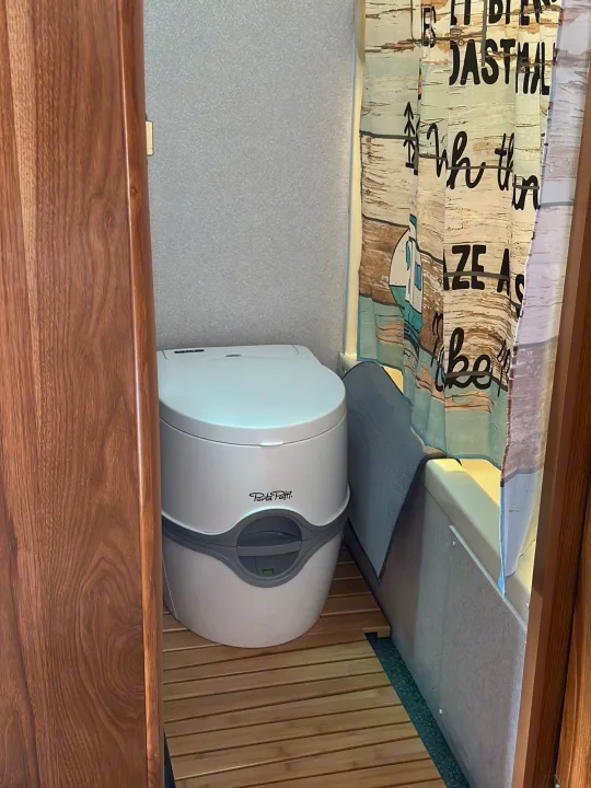 RV rental bathroom features a portable toilet (Porta Potti), slatted wooden floor mat, and a shower with a themed curtain.. Forest River Cherokee Lite 2000