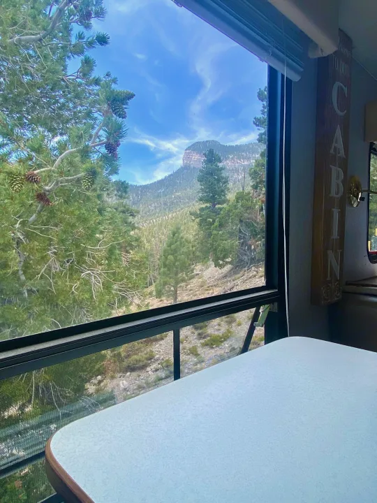 RV rental view: Lush pine trees and distant mountains through the window. A white table and 'CABIN' sign complete this cozy escape.. Forest River Cherokee Lite 2000
