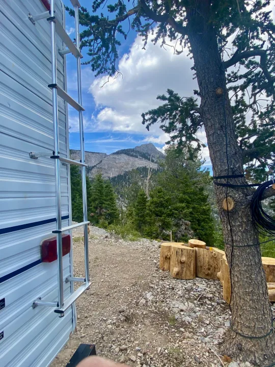 Back of an RV rental parked in a scenic mountain setting with a large tree and log seating.. Forest River Cherokee Lite 2000