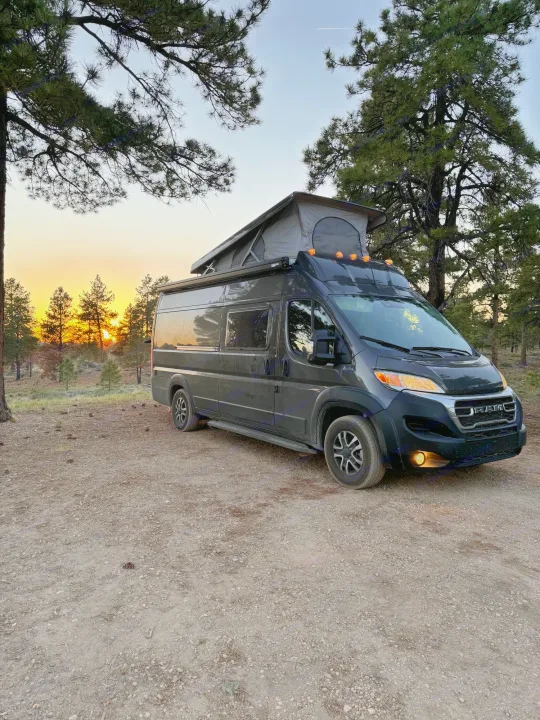 A dark gray rv rental camper van with a pop-top roof, parked in a pine forest at sunset. Its lights glow.. Noovo Pop 2024