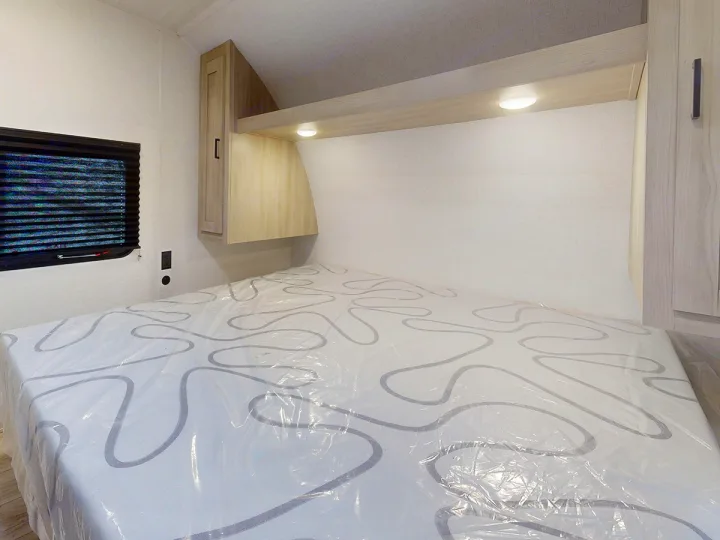 RV rental bedroom featuring a bed with a plastic-covered mattress, overhead cabinets, and a window with blinds.. East to West Silver Lake 3250QB 2025
