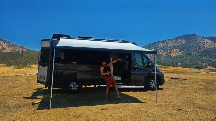 Enjoying an RV rental: A person poses by a dark grey camper van with extended awning, in a sunny, dry, mountainous landscape.. Entegra Coach Ethos 2024