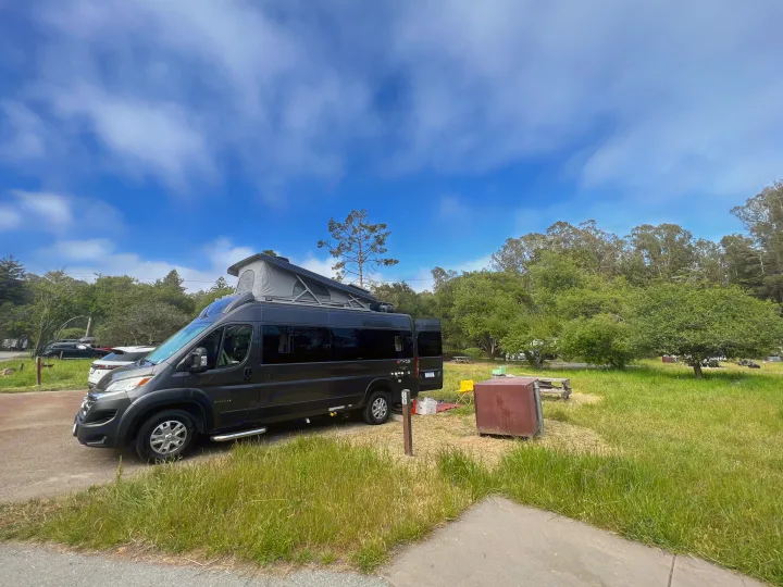 Dark grey camper van RV rental with pop-top roof extended at a sunny campsite amidst green grass & trees.. Entegra Coach Ethos 2024
