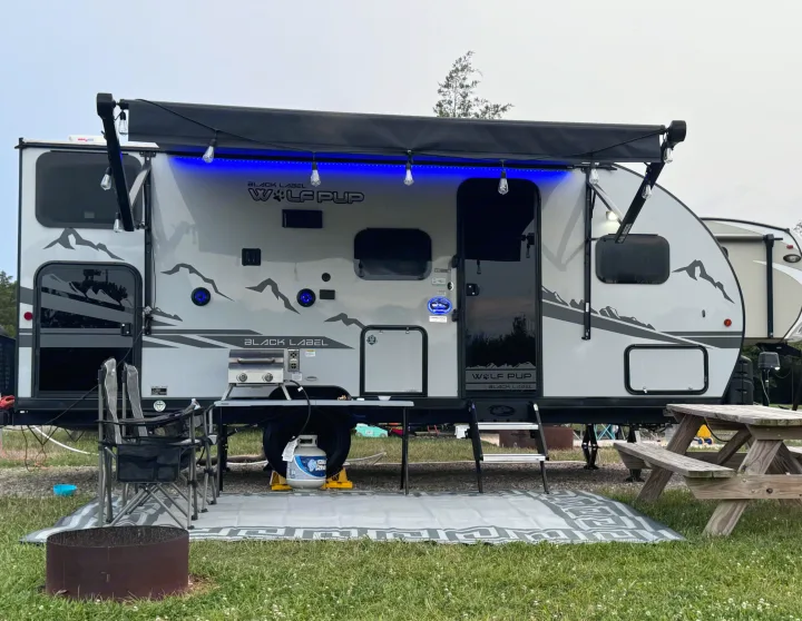 Grey Wolf Pup rv rental at a campsite, featuring blue awning lights, grill, chairs, and outdoor mat for cozy camping.. Forest River Cherokee Wolf Pup Black Label 2022