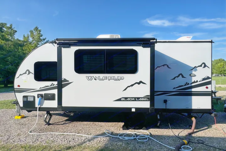 Light gray Wolf Pup Black Label travel trailer, set up with water/power at a sunny campsite. Ideal for your RV rental getaway.. Forest River Cherokee Wolf Pup Black Label 2022