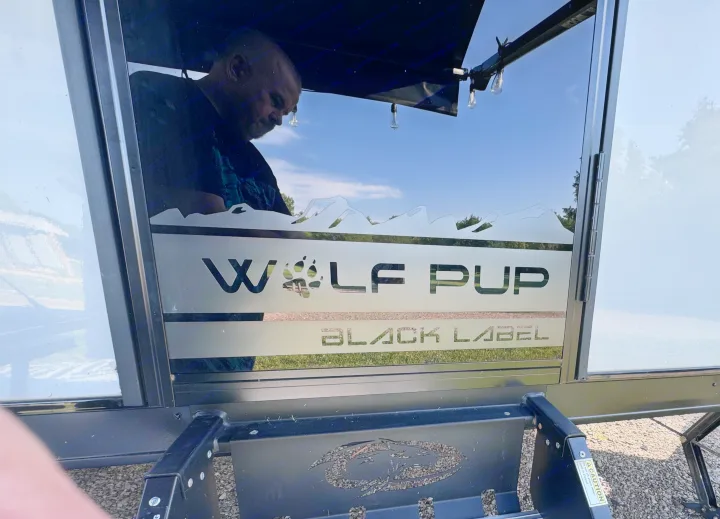 Side of an RV rental: a window with etched 'Wolf Pup Black Label' branding, paw print, and mountain design. A person is reflected.. Forest River Cherokee Wolf Pup Black Label 2022