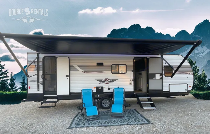 Shasta RV rental with awning extended, open doors, chairs, and mat. Mountain backdrop on a gravel site.. Shasta 26DB 2022