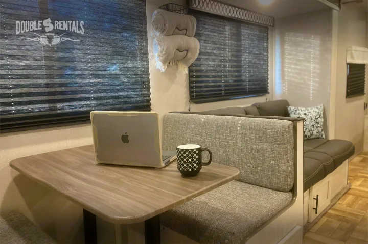 RV rental interior: dinette with laptop & mug, cozy bench seating & sofa, window blinds, and rolled blankets.. Shasta 26DB 2022