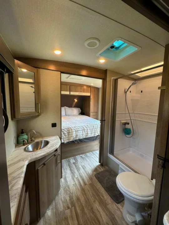 This RV rental interior features a compact bathroom with a sink, shower, and toilet, leading into the cozy bedroom.. Grand Design Imagine 2022