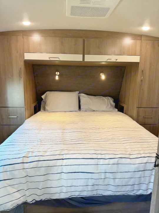 Cozy RV rental bedroom with striped bedding, two pillows, overhead cabinets, and wall-mounted lights.. Grand Design Imagine 2022
