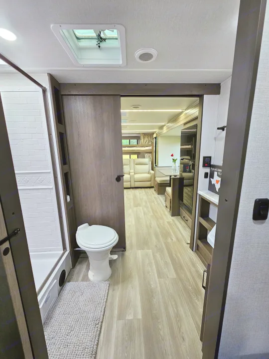 RV rental interior: A bright bathroom with toilet and shower, opening to a modern hallway and living area with light wood floors.. Grand Design Transcend Xplor 2024