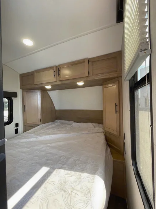 RV rental bedroom with a large bed, white quilt, overhead cabinets, side wardrobes, and sunny window.. Forest River Cherokee Wolf Pup 2023