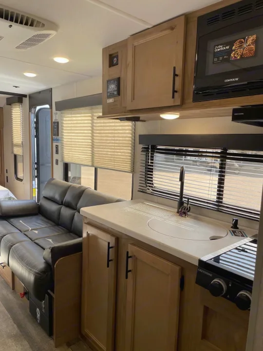 Inside an RV rental: a sleek kitchen with a Greystone stove & Contoure microwave, plus a spacious black sofa and large windows.. Forest River Cherokee Wolf Pup 2023