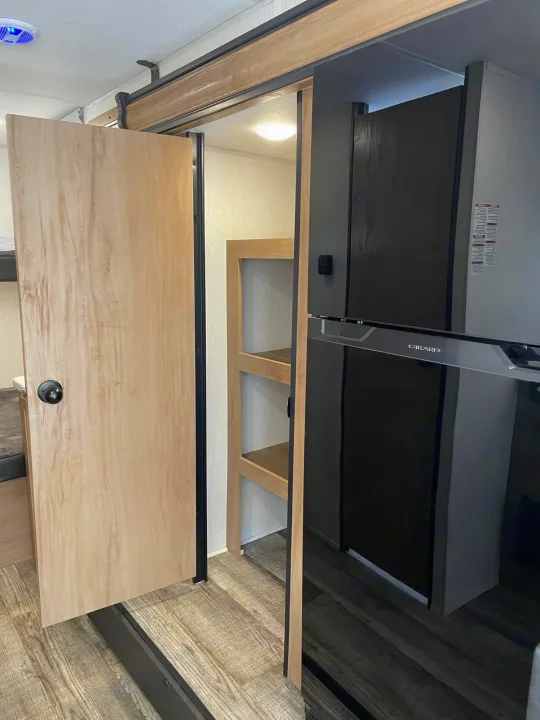 This rv rental interior features a light wood pocket door to bunks, shelving, & a modern black Girard refrigerator.. Forest River Cherokee Wolf Pup 2023