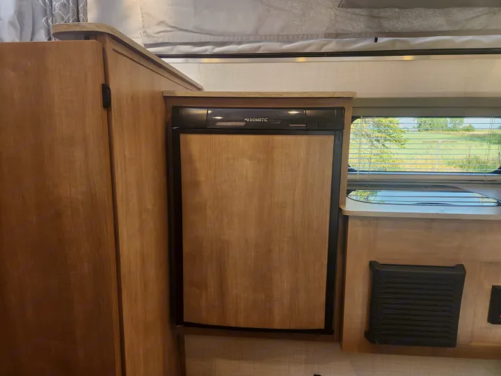 RV rental interior: Dometic wood-paneled fridge, light wood cabinets, and a window overlooking green fields.. Palomino SS1500 2023