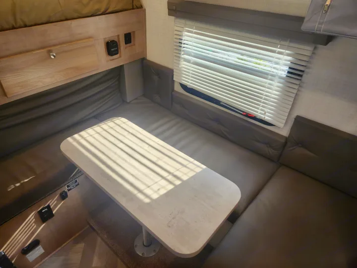 Bright RV rental interior: a comfortable dinette with light wood table, dark benches, and sunlit window blinds.. Palomino SS1500 2023
