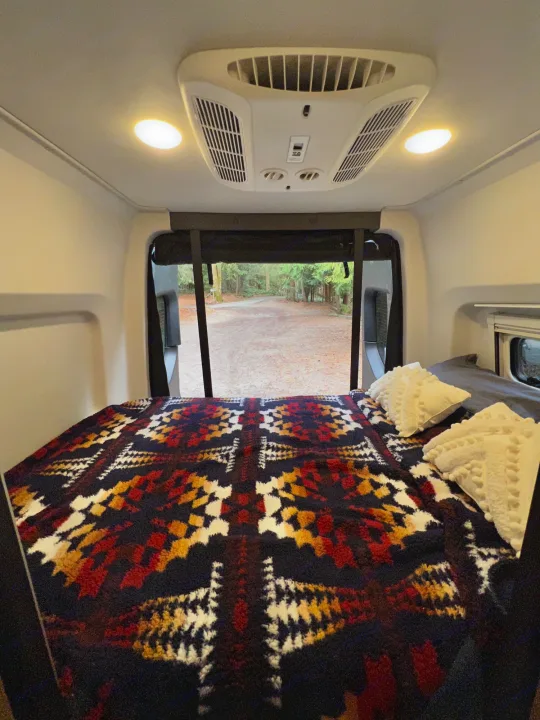 Cozy RV rental bed with a vibrant blanket and pillows. The open back doors reveal a peaceful road and trees outside.. Mercedes Sprinter Tiffin GH1 2024