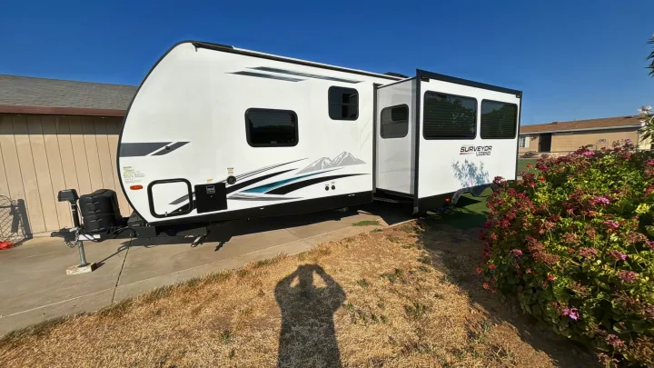White Surveyor Legend travel trailer for RV rental, parked on a driveway with its side slide-out extended, under a clear sky.. Forest River Surveyor Legend 2023