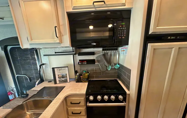 Compact RV rental kitchen with overhead microwave, a stove/oven, sink, and light wood cabinets, designed for road trips.. Jayco Greyhawk 2023