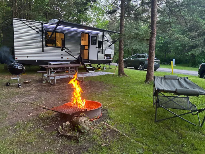 An RV rental is set up at a wooded campsite with a roaring campfire, grill, and picnic area, perfect for a relaxing getaway.. East to West Silver Lake 2023