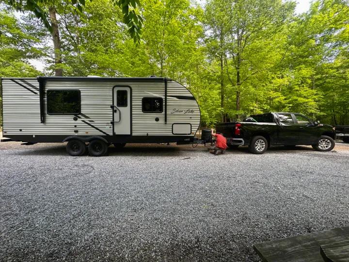 Person connects an RV rental travel trailer to a truck on a gravel campsite, surrounded by vibrant green trees.. East to West Silver Lake 2023